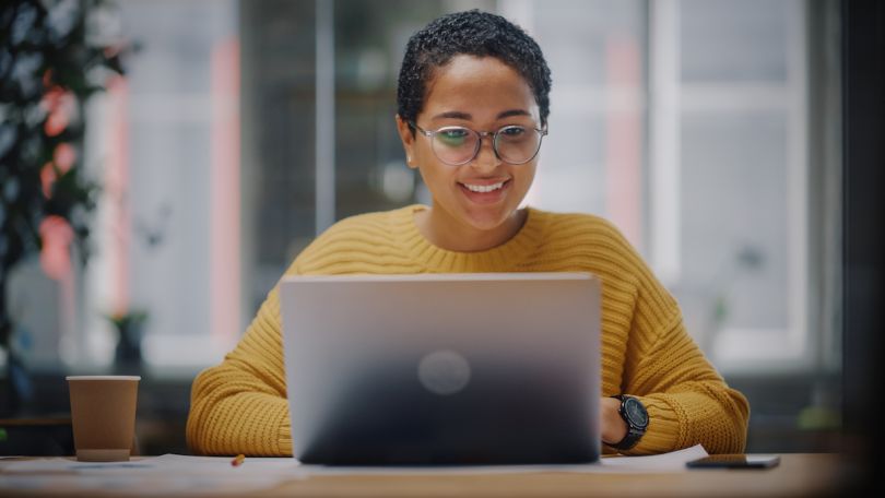 A woman in a yellow sweater smiles at a laptop screen