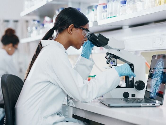 A cancer researcher in a laboratory looks at a specimen through a microscope while comparing their findings to an image displayed on a laptop screen.&nbsp;