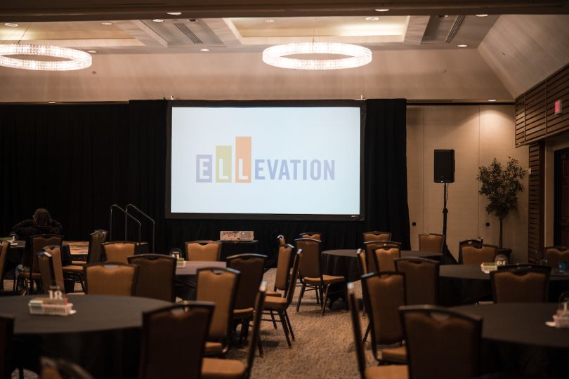 Empty ballroom with circular tables, chairs and screen at back with Ellevation logo