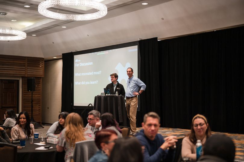 Indoor ballroom set up with presentation screen and podium, people discussing topic onscreen