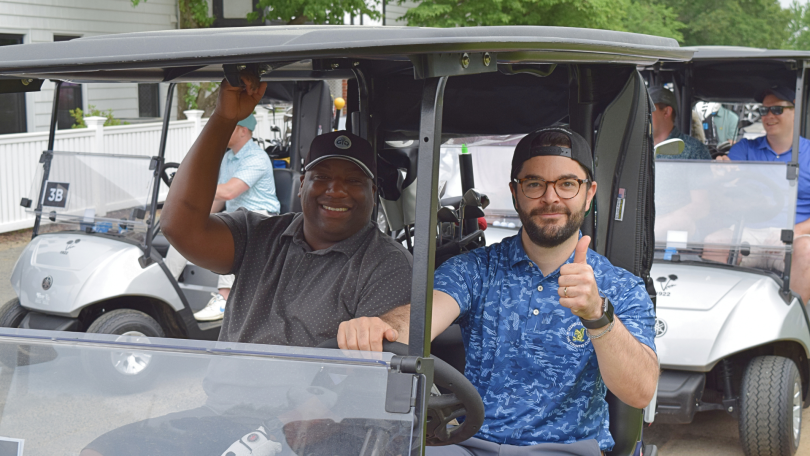 CarGurus team members smile and give a thumbs-up while driving a golf cart at a charity event.&nbsp;