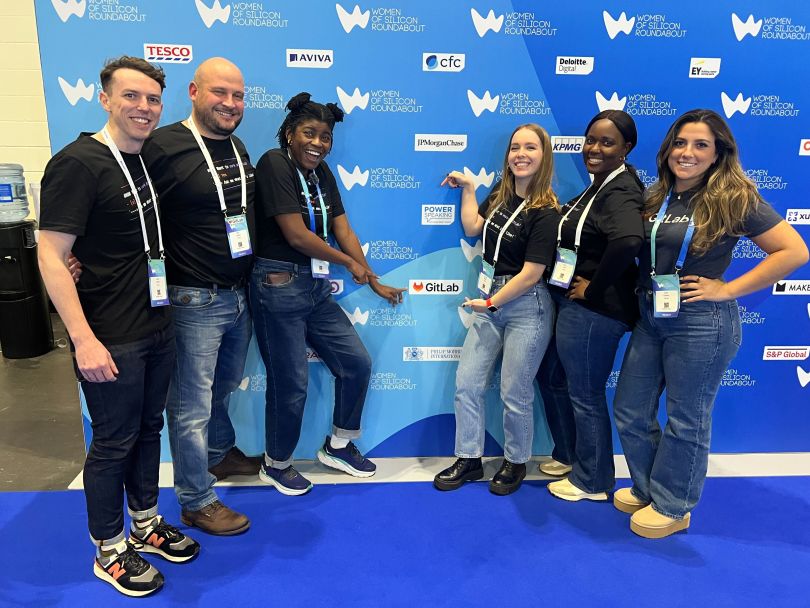 Group photo of GitLab team members in front of a step-and-repeat backdrop at a GitLab-sponsored event, pointing to the GitLab logo on the backdrop.&nbsp;