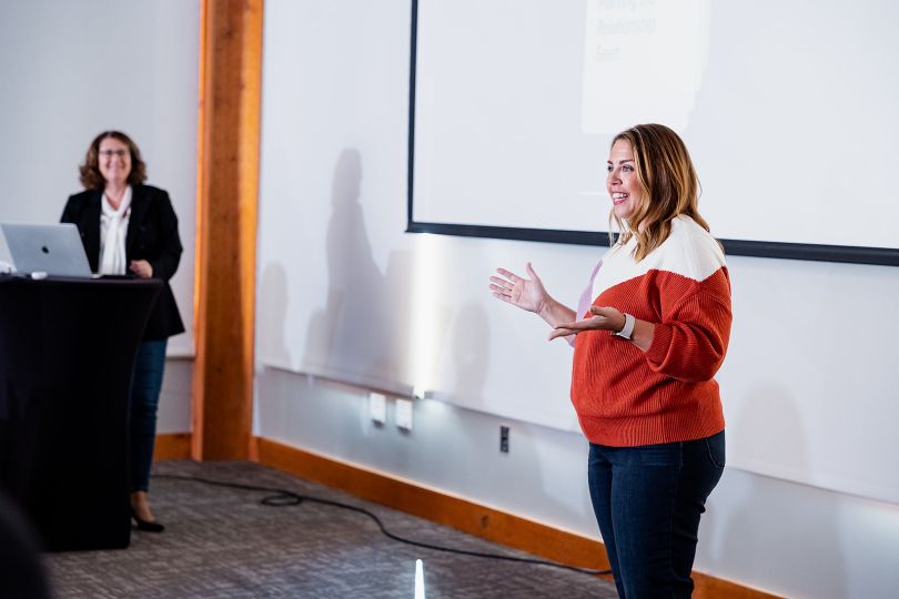 A leader stands at the front of a room, smiling and addressing a team, while Jennifer Guisbert stands, smiling attentively, running the presentation on a laptop at the lectern nearby.&nbsp;