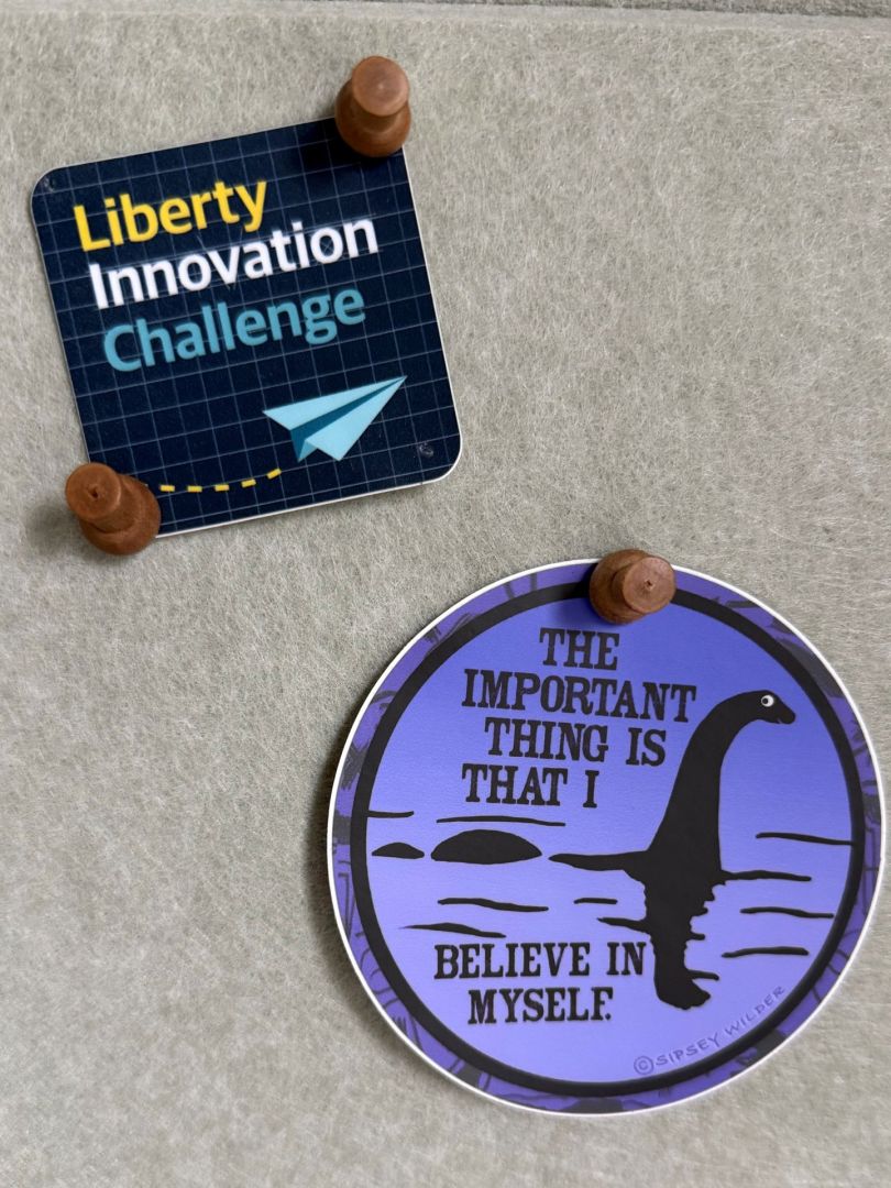A photo of two stickers pinned against a felt board, with one bearing the words “Liberty Innovation Challenge” and another bearing the image of the Loch Ness monster and the statement, “The most important thing is that I believe in myself”