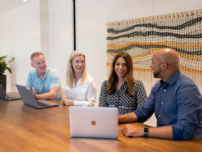 Telesign teammates sit next to each other at a table at the company’s 2024 Revenue Kickoff event, looking at laptops together and smiling.&nbsp;