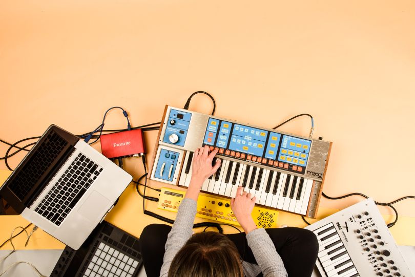Overhead view of person sitting cross-legged on the floor playing a keyboard and surrounded by a laptop and other sound equipment.&nbsp;