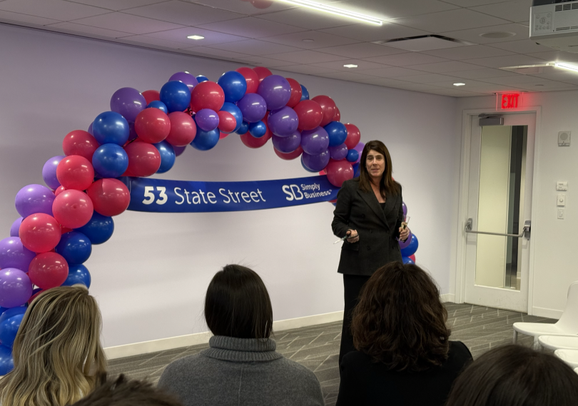  A Simply Business leader stands in front of a balloon archway while speaking in front of employees during a company event