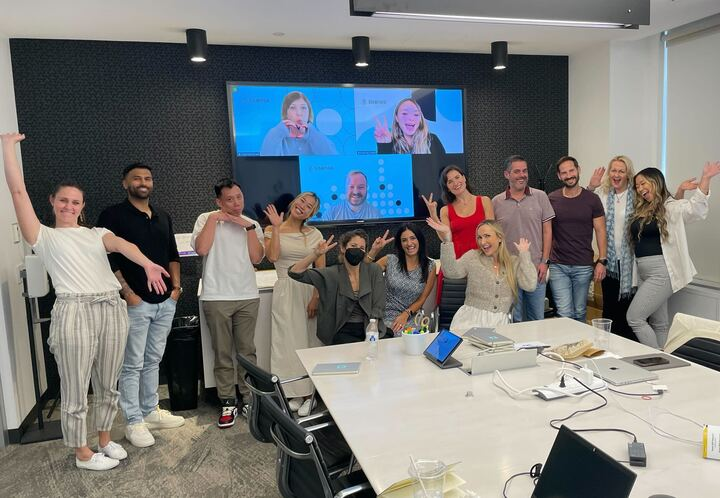 12 Sisense team members do silly poses for a team photo in an office conference room as three team members pose behind them over Zoom