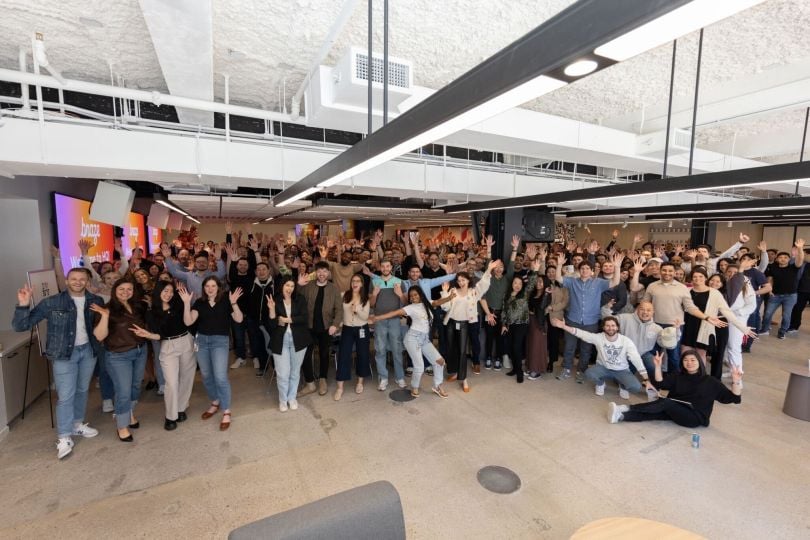 Braze’s team poses for a large group photo in a spacious, industrial-decor office space with large screens featuring Braze’s logo in the background.