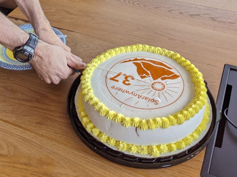 A cake reading SolarAnywhere 3.7 with a logo of mountains and a sunburst is cut by a person wearing a watch and holding the knife with both hands.