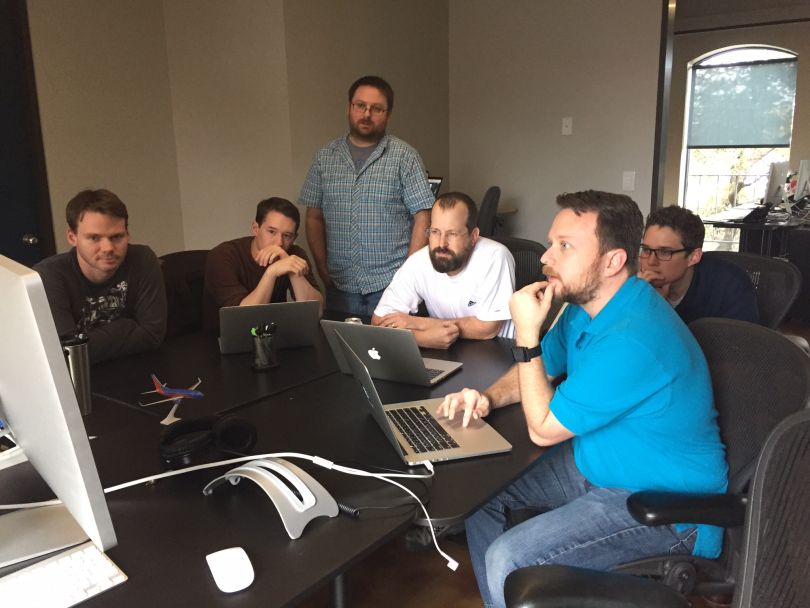 A group of ForeFlight employees gather around a table with three laptops among them and focus on a large secondary monitor.
