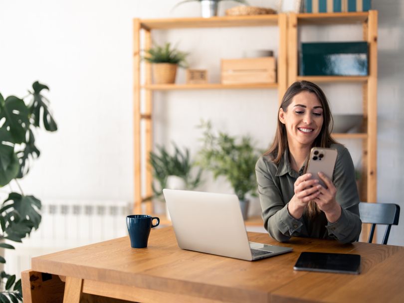 A smiling woman checks her finances on her phone while working in her home office
