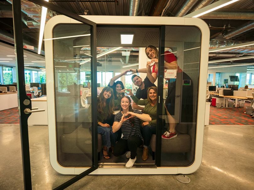 Restaurant365 employees sit in group inside a square meeting pod in their office, forming heart shapes with their hands.