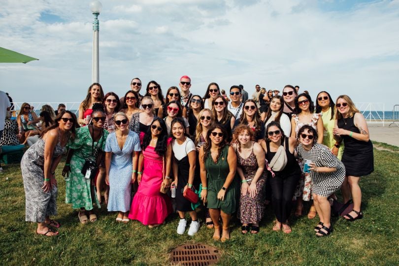 Sprout employees in summer clothes post for a large group photo on a green area in front of a large body of water.