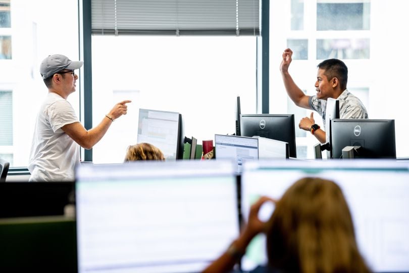 Tango Card employees talk animatedly over the top of their monitors at two standing desks.