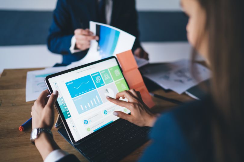 A woman sits at a conference table consulting an analytic dashboard on her tablet