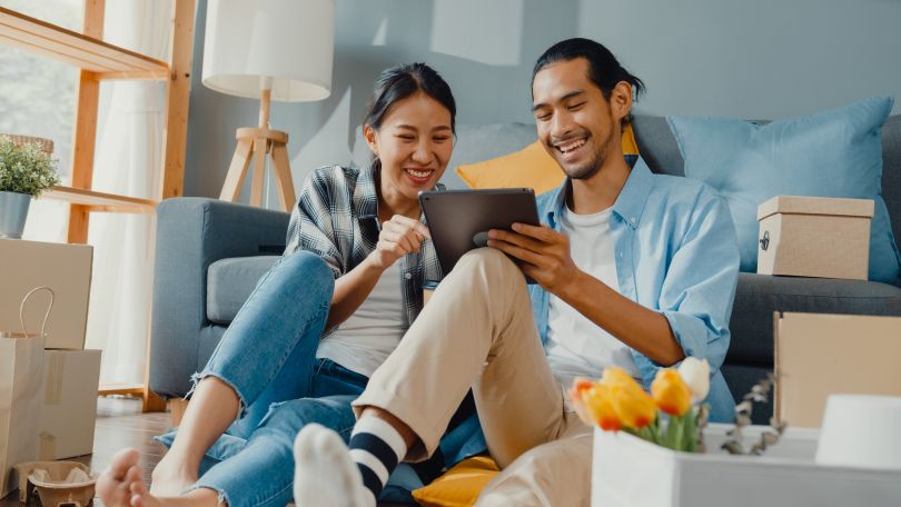A smiling couple sits among moving boxes and consults a tablet.
