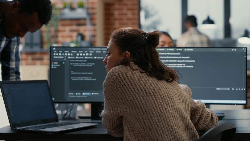 A software developer working at a desk talking to a colleague