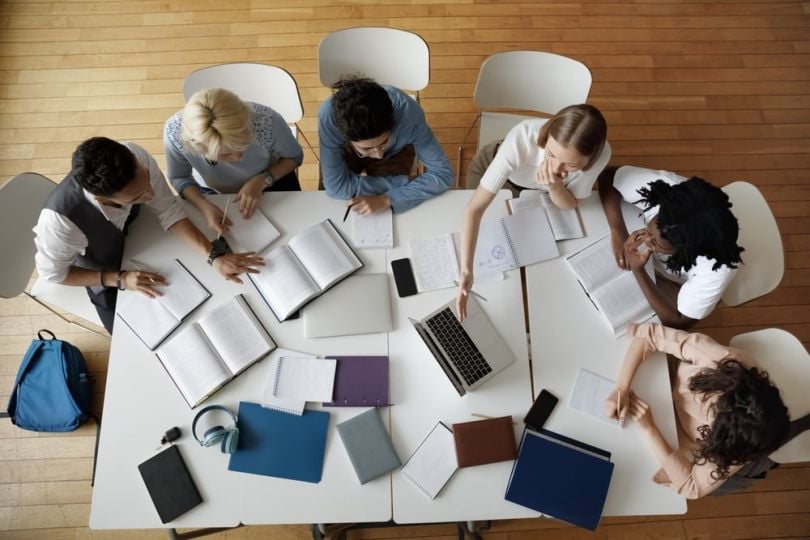 In-office workday meeting, overhead view of coworkers meeting at a table with laptops and papers. 