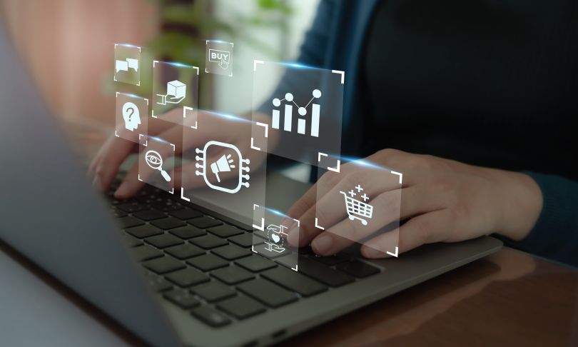 A person is using a laptop with graphical icons representing online activities like shopping and data analysis floating above their hands, indicating digital marketing.