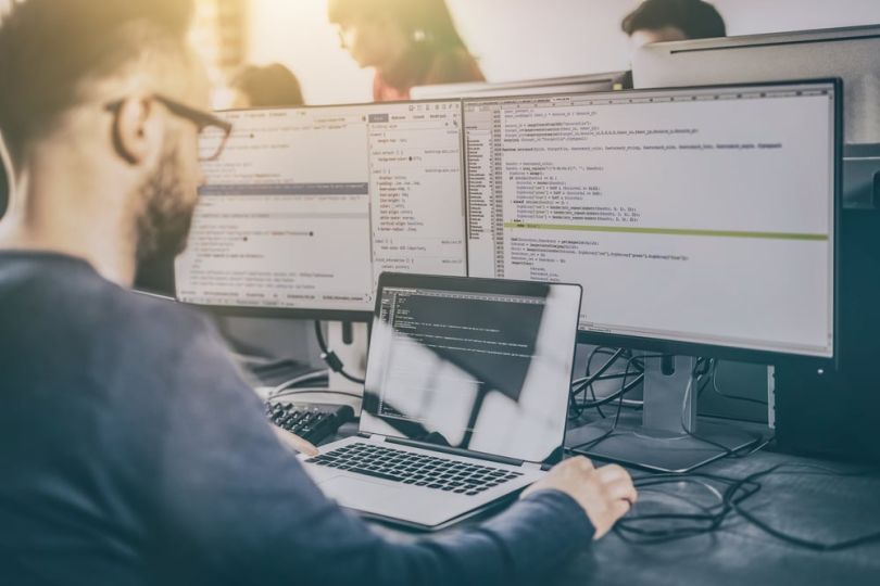 A developer writing code at a desk with three computer monitors.