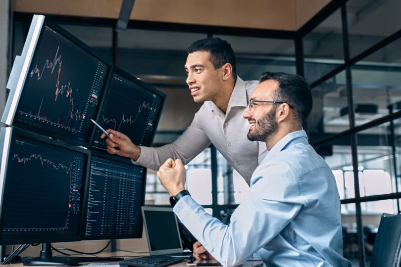 Two professionals looks at trading stats on a screen.&nbsp;