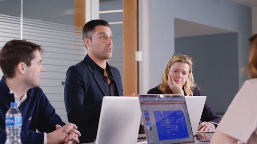  Candid photo of Zak Dabbas speaking to team members in a meeting with computers on the table.