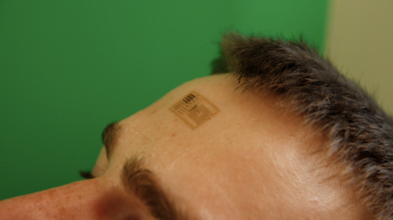 electronic tattoo, shown as thin patch-like material, placed on person's forehead.